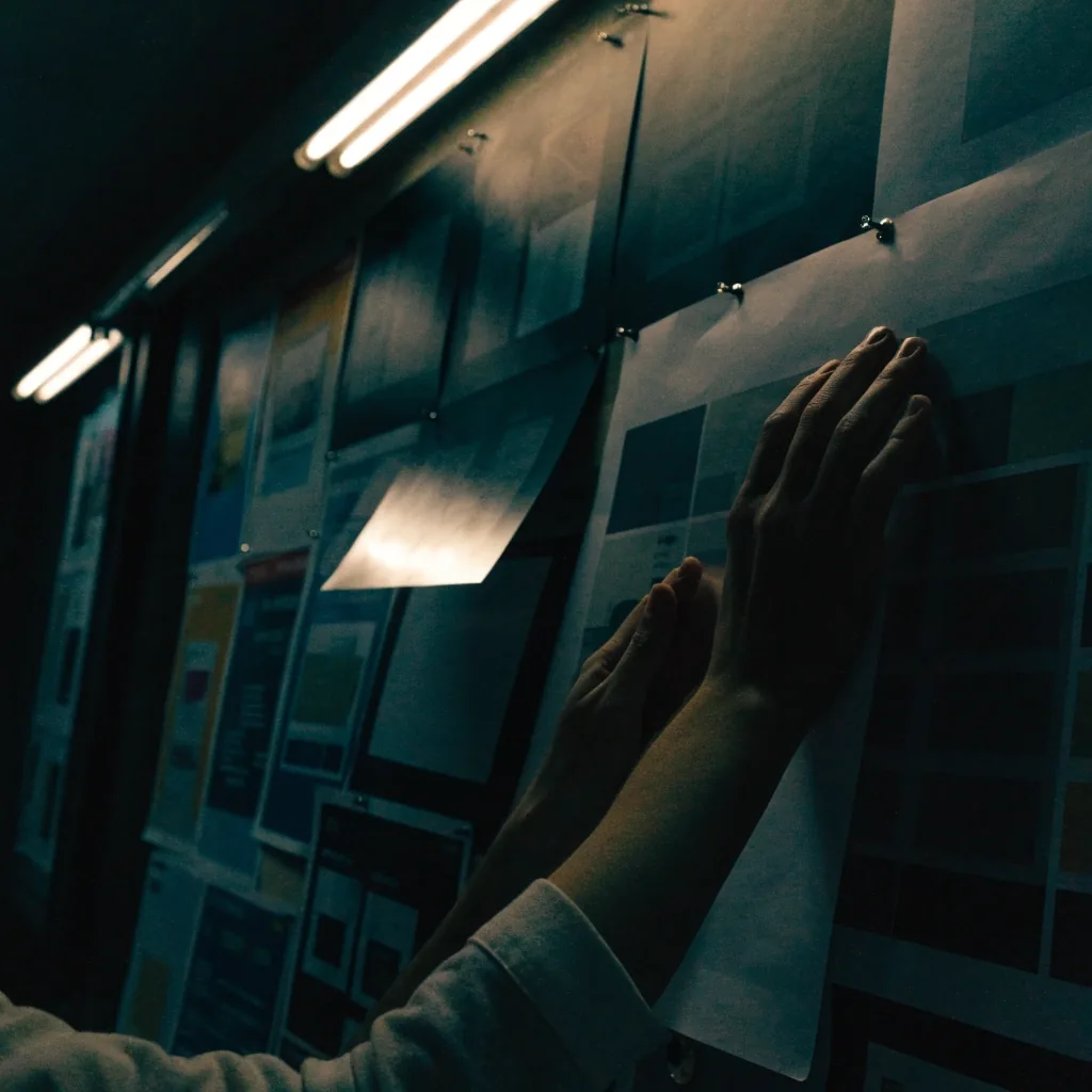 Hands pinning a printed interface frame to a wall of artboards in a dim studio